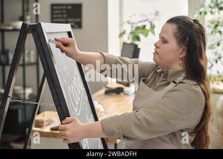 Young female worker of cafe with mental disability writing down menu with white chalk on blackboard while standing in front of camera Stock Photo