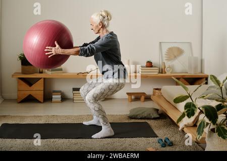 Side view of aged active woman in sportswear holding fitball in front of herself while doing squats on the floor of bedroom in the morning Stock Photo