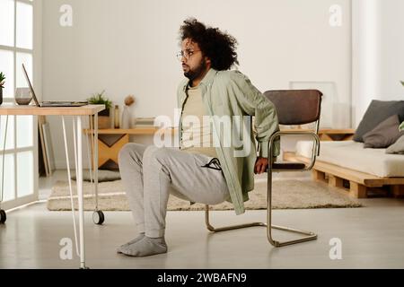 Multiethnic young man in casualwear doing squats in front of laptop while holding by chair and looking at screen during physical exercise Stock Photo