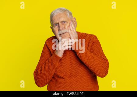 Dental problems. Senior old man touching cheek, closing eyes with expression of terrible suffer from painful toothache, sensitive teeth, cavities. Elderly grandfather pensioner on yellow background Stock Photo