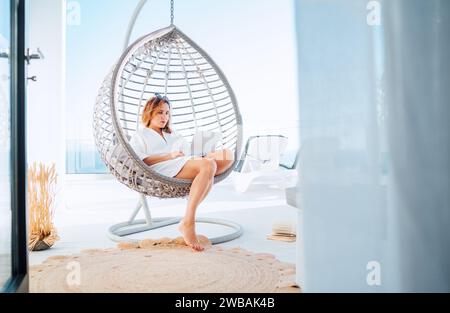 Beautiful middle-aged woman sitting in a comfortable hanging chair on open house terrace with a modern laptop working remotely at home office. Modern Stock Photo