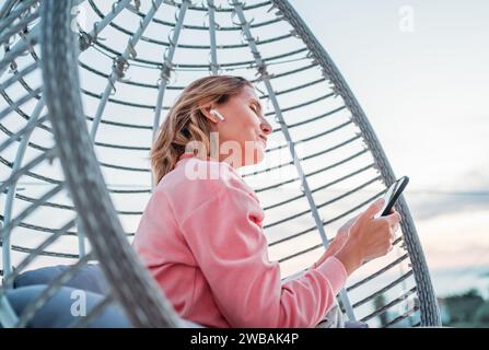 Beautiful middle-aged woman listening Earbuds smartphone music sitting in comfortable hanging chair on open house terrace Stock Photo