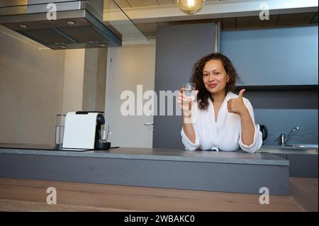 Beautiful brunette, curly young woman smiles, thumbs up looking at camera, holds a glass of mineral water, standing at counter in modern minimalist ki Stock Photo