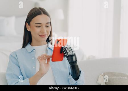 Smiling young woman with prosthetic arm using smartphone, experiencing accessibility technology at home Stock Photo