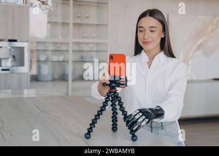 Smiling woman with a prosthetic arm setting up a mobile phone on a tripod for a video call in a modern kitchen. Stock Photo