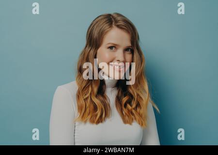 Charming woman with curly hair and white turtleneck smiling gently, exuding warmth and style against a blue background Stock Photo