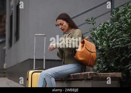 Being late. Worried woman with suitcase looking at watch on bench outdoors Stock Photo
