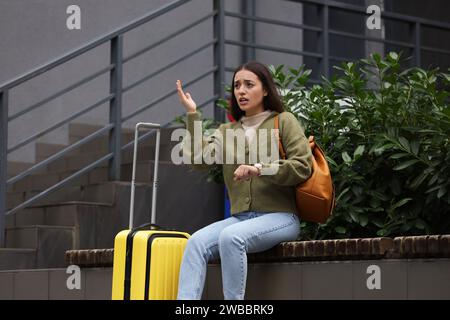 Being late. Worried woman with suitcase sitting on bench outdoors Stock Photo