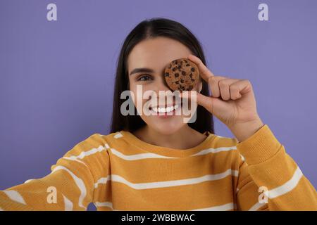 Young woman with chocolate chip cookie taking selfie on purple background Stock Photo