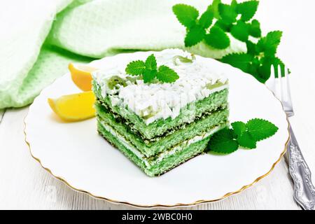 Piece of cake with mint and orange, white cream, confit, green sponge cake, sprinkled with white chocolate and green jelly in a plate, towel on wooden Stock Photo