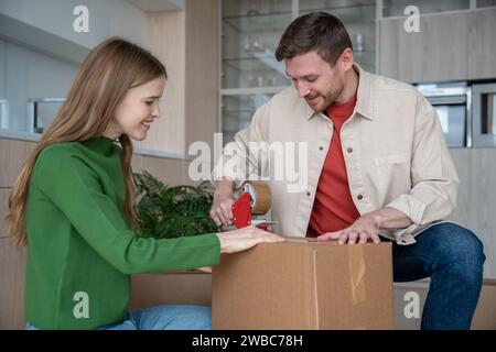 Cheerful couple packing cardboard boxes using sticky tape prepare to move in new apartment together. Stock Photo