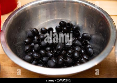 Pitted black olives glistening with moisture, contained in a reflective metal bowl set on a wooden kitchen counter. Stock Photo