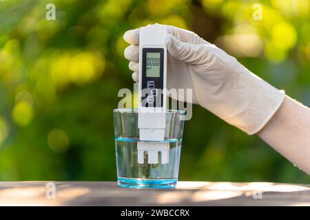 pH meter in hands with gloves, glass of water on blurred background of nature. Measurement of characteristics of drinking water. hardness of water. pu Stock Photo