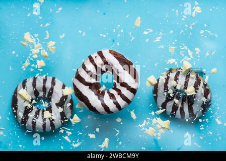 Three chocolate covered donuts with white chocolate chips falling on a pastel blue background. Close up, with copy space Stock Photo
