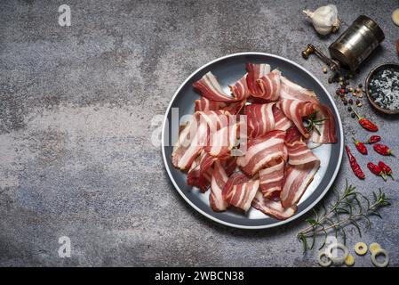 Rolled smoked bacon in a plate on a light gray background. Various spices are around - garlic, rosemary, pepper, red hot peppers and a pepper mill Stock Photo
