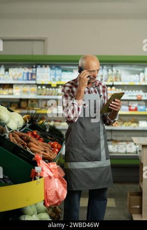 Old male sales assistant in a grocery using digital tablet and talking on cell phone. Owner with digital tablet preparing online grocery order for his Stock Photo