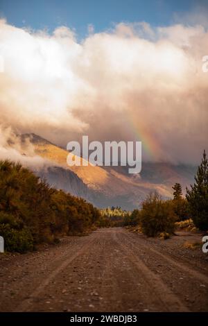 A country road surrounded by lush trees in mountains, with rainbow in the background Stock Photo
