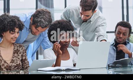 Happy young female employee discussing online project, showing computer presentation to skilled team leader in eyeglasses. Friendly diverse colleagues Stock Photo