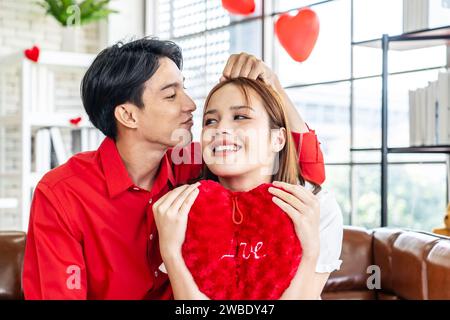 Portrait of asian young couple sitting on sofa smiling and holding red heart pillow in living room for romance and celebrating valentine day. Stock Photo