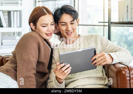 Happy asian young couple looking at digital computer tablet screen, relaxing on comfortable couch. Loving couple shopping online in internet store Stock Photo