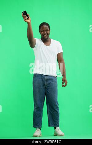 A man in a white T-shirt on a green background raised his phone up. Stock Photo