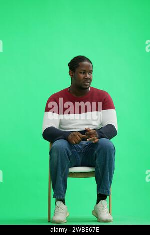 A man in a multi-colored jacket with a backpack on a green background sits on a chair Stock Photo
