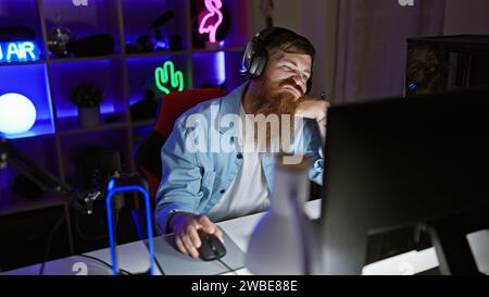 Bored young redhead man streamer getting tired, struggling to stay awake playing video game, engulfed in dark night in his gaming room Stock Photo