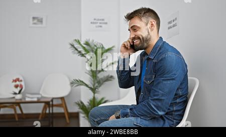 Handsome hispanic man entranced in lively phone talk, sitting relaxed on chair in waiting room, beaming a bright smile while navigating his smartphone Stock Photo