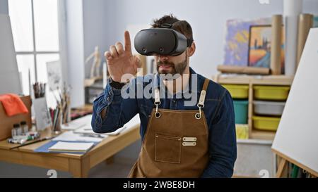 Young, bearded hispanic artist in apron manically draws on canvas in art studio using futuristic virtual reality glasses Stock Photo