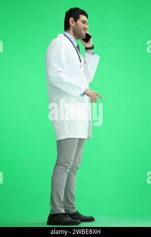Male doctor on a green background in full growth talking on the phone, in profile Stock Photo