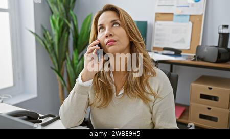 A thoughtful hispanic woman converses on a smartphone in a modern office setting, exemplifying professionalism and engagement. Stock Photo