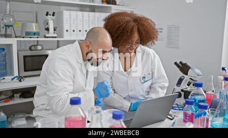 Two amiable scientists happily speaking and discovering breakthroughs together on laptop in busy laboratory amidst test tubes and microscopes Stock Photo