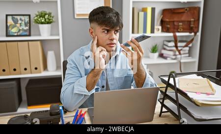 Handsome young hispanic man working diligently in office, using laptop and sending voice messages via smartphone - serious commitment to success Stock Photo