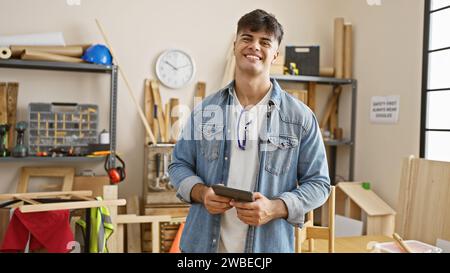 Smiling young hispanic man, a professional carpenter, engrossed in his carpentry work indoors, skillfully using a touchpad in his well-furnished works Stock Photo