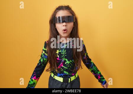 Adorable hispanic girl's shocked expression, wearing vr glasses, open-mouthed in disbelief and fear on isolated yellow background Stock Photo