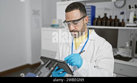 A bearded man in labcoat and safety glasses works attentively on a tablet in a modern laboratory. Stock Photo