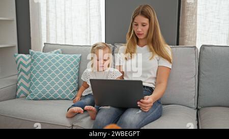 Serious caucasian mother and little daughter engrossed, chilling on their living room sofa at home, completely absorbed in watching an intriguing vide Stock Photo