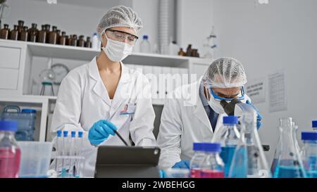 Two scientists, in full medical gear, analyzing covid19 samples at the lab, scribbling notes on touchpad under the microscope's zoom Stock Photo