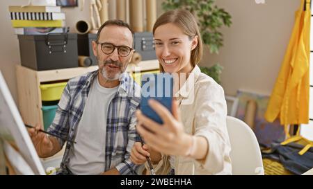 Two smiling artists immersed in taking a selfie with smartphone amidst the colorful chaos of their art studio Stock Photo