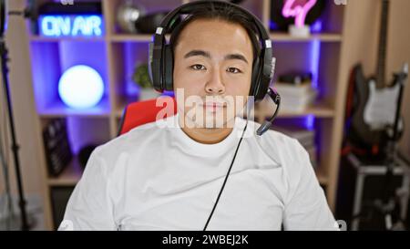 Intense night stream, young chinese man, a committed streamer, with serious gaze, plugged into gaming room, headset ready, engrossed in futuristic cyb Stock Photo