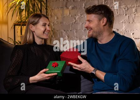 romantic couple exchanging with surprise gifts while sitting on sofa at home. valentine's day or wedding anniversary present Stock Photo