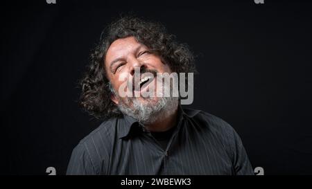 Portrait of latin man with white beard and black curly hair with funny expression, looking up, wearing black shirt against black background Stock Photo