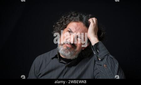 Portrait of latin man with white beard and black curly hair with confused expression, scratching head with his hand, wearing black shirt against black Stock Photo