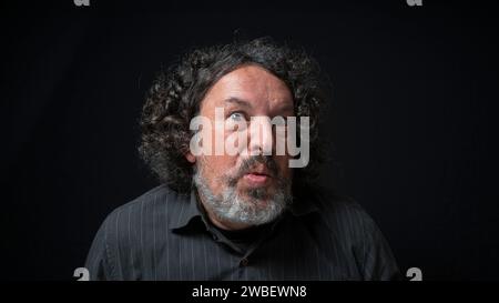 Portrait of latin man with white beard and black curly hair with funny expression, grimacing with eyes and mouth, wearing black shirt against black ba Stock Photo
