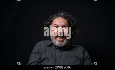 Portrait of latin man with white beard and black curly hair with happy and excited expression, wearing black shirt against black background Stock Photo