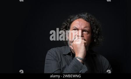 Portrait of latin man with white beard and black curly hair with doubtful and angry expression, with his hand on his face, wearing black shirt against Stock Photo