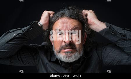 Portrait of latin man with white beard and black curly hair with very angry expression, grabbing his hair with hands, wearing black shirt against blac Stock Photo