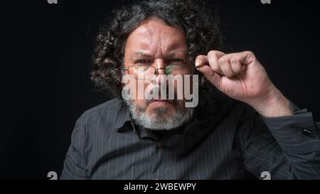 Portrait of latin man with white beard and black curly hair with curious expression, looking at camera suspiciously, wearing black shirt against black Stock Photo