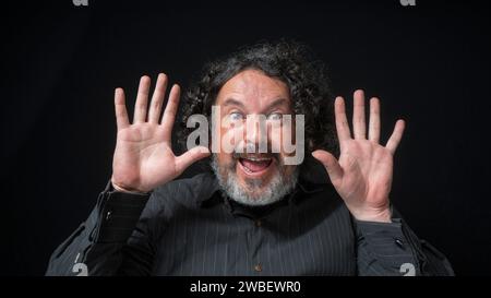 Portrait of latin man with white beard and black curly hair with happy and excited expression, smiling looking at camera, wearing black shirt against Stock Photo