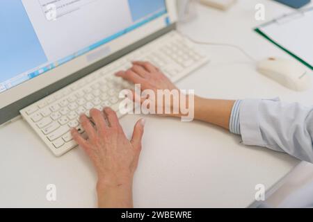 Close-up top view hands of unrecognizable female doctor in white coat working and typing on laptop computer keyboard at medical office in clinic. Stock Photo